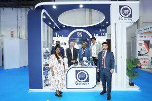 A professional group photo of six ORP Germany team members posing in front of their sleek white and blue trade show stand. The booth prominently displays the ORP Germany logo and various automotive spare parts, including filters and engine components, neatly arranged on the counters and shelves.